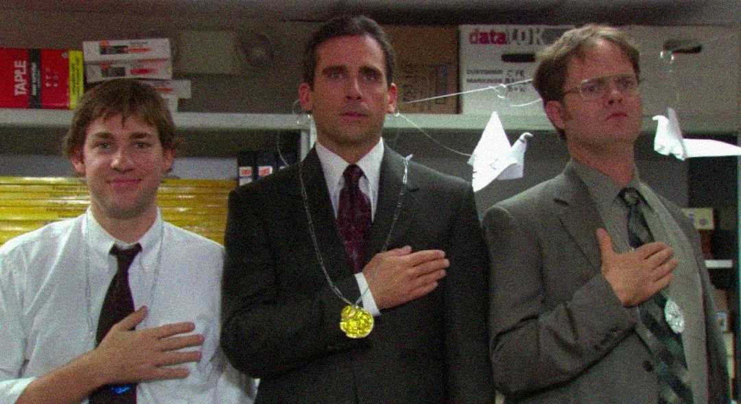 Three men in suits stand indoors with their right hands over their hearts, wearing gold medals on strings around their necks. The background shows office supplies and two white origami birds hanging.