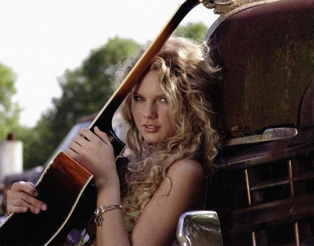 A young woman with long, curly blonde hair holds an acoustic guitar, leaning against an old vehicle outdoors on a sunny day. Trees are visible in the blurred background.