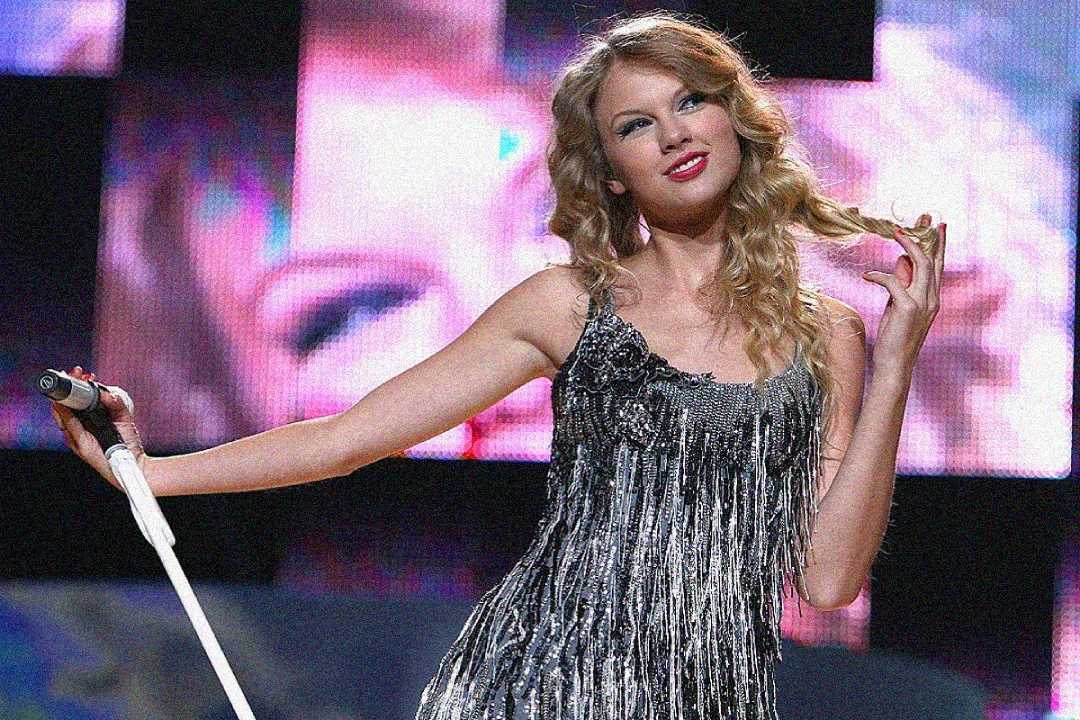 A woman in a sparkly, fringed dress holds a microphone stand and twirls her hair, performing on stage with a large screen displaying her image in the background.