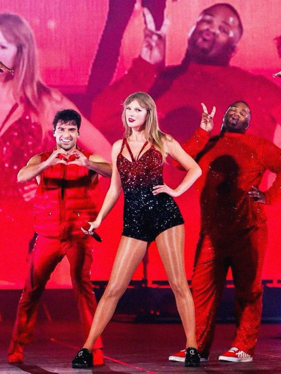 A woman in a sparkling red and black outfit stands center stage with two male dancers in red, one making a heart shape with his hands and the other giving a peace sign, against a bright pink and red background.