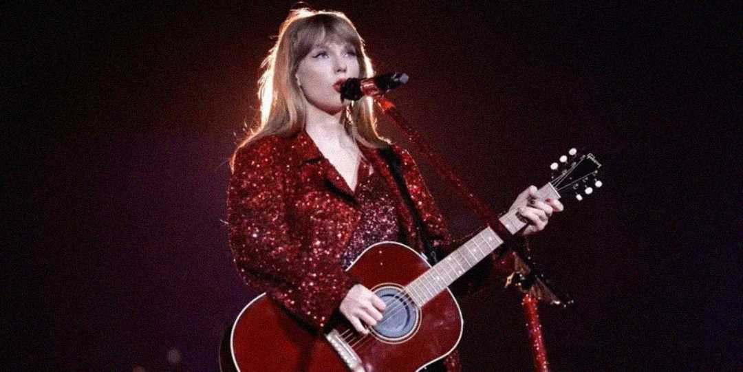 A woman in a sparkling red outfit plays a red acoustic guitar and sings into a microphone on stage, illuminated by a bright light from behind.