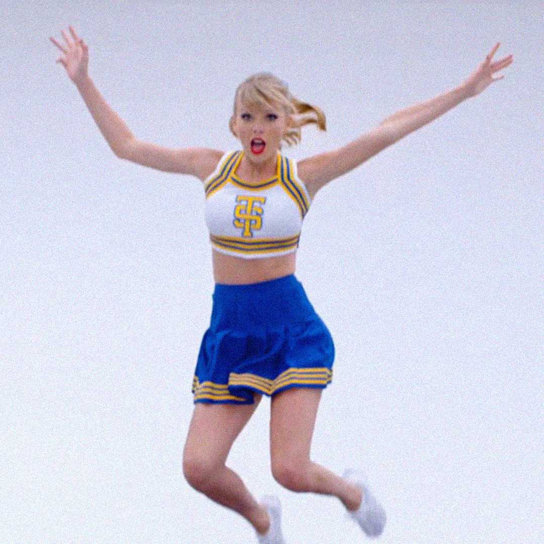 A woman in a cheerleader outfit with a white and yellow crop top and blue skirt jumps in the air with arms raised and mouth open, against a plain light background.