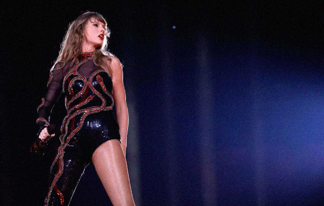 A woman in a sparkling black and red outfit performs on stage under dramatic lighting, holding a microphone and looking confidently to the side.