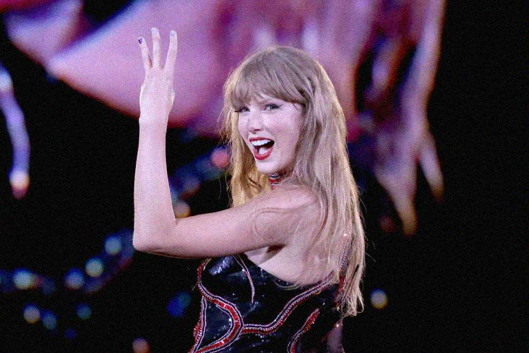 A woman with long blonde hair, wearing a sparkly black and red outfit, smiles and holds up three fingers while performing on stage with vibrant lights in the background.