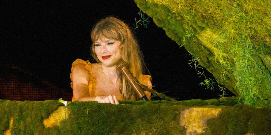 A woman in a yellow ruffled dress with long blonde hair stands behind a piano covered in green moss, smiling softly under warm stage lighting.