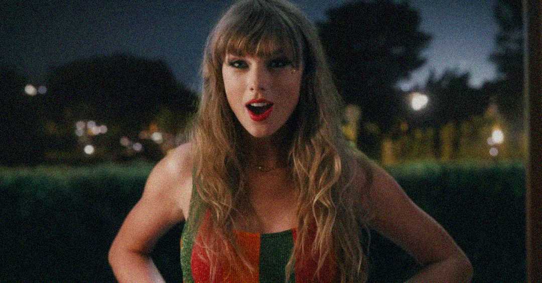 A woman with long wavy hair and bangs stands outdoors at night, wearing a sleeveless top. She has a surprised expression with her mouth open and is looking toward the camera. Trees and lights are visible in the background.