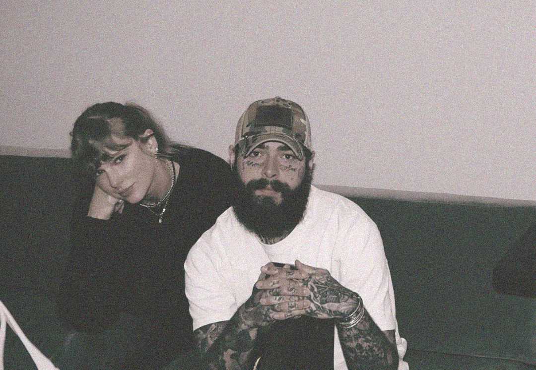 A woman with long hair rests her head on her hand while sitting next to a bearded man with tattoos, wearing a cap and a white shirt. Both are seated on a couch against a plain wall.