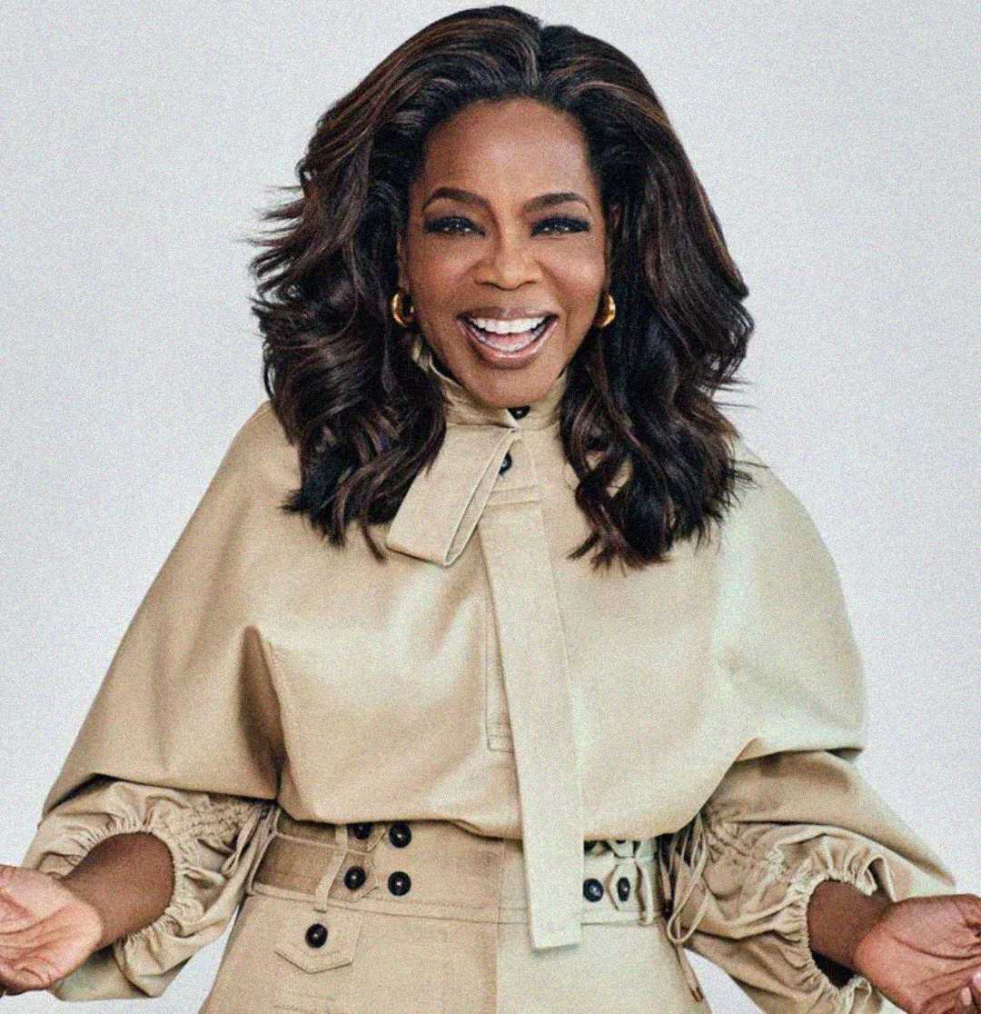 A woman with wavy dark hair and gold hoop earrings smiles warmly with open arms. She is wearing a light beige belted outfit with a high neck and button details, standing against a plain background.