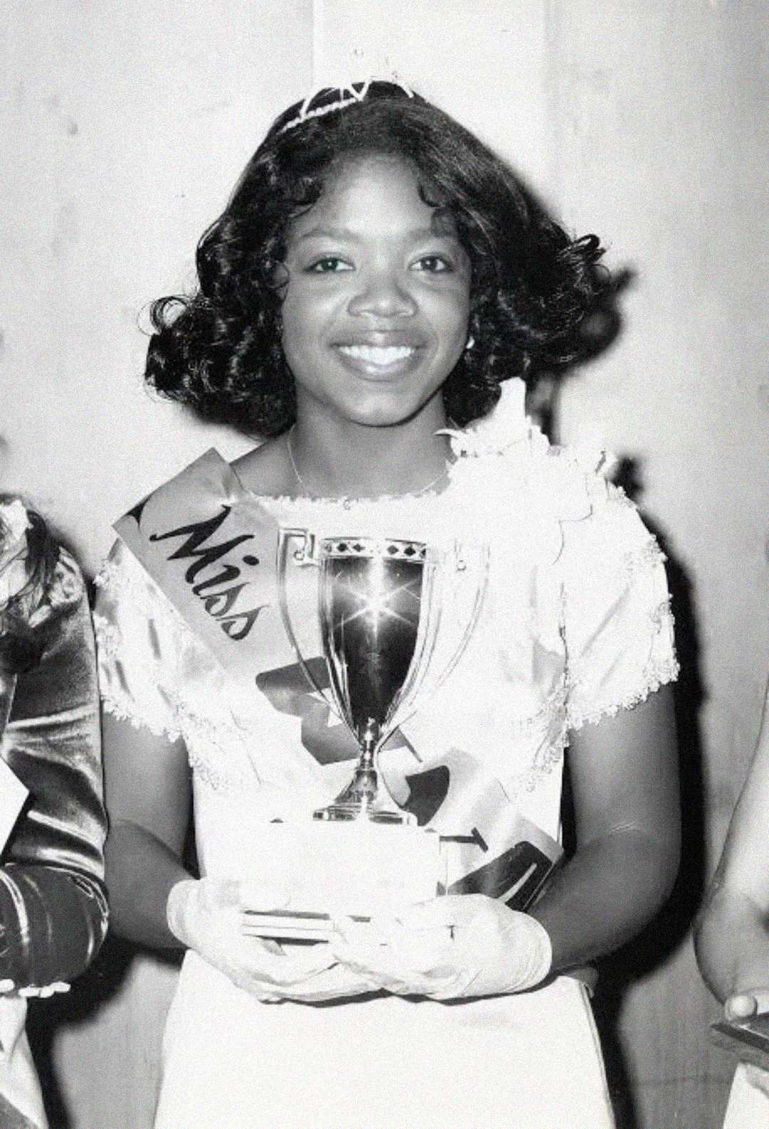 A young woman wearing a sash that reads "Miss" and a tiara smiles while holding a trophy. She is dressed in a formal gown and gloves, standing against a plain background.