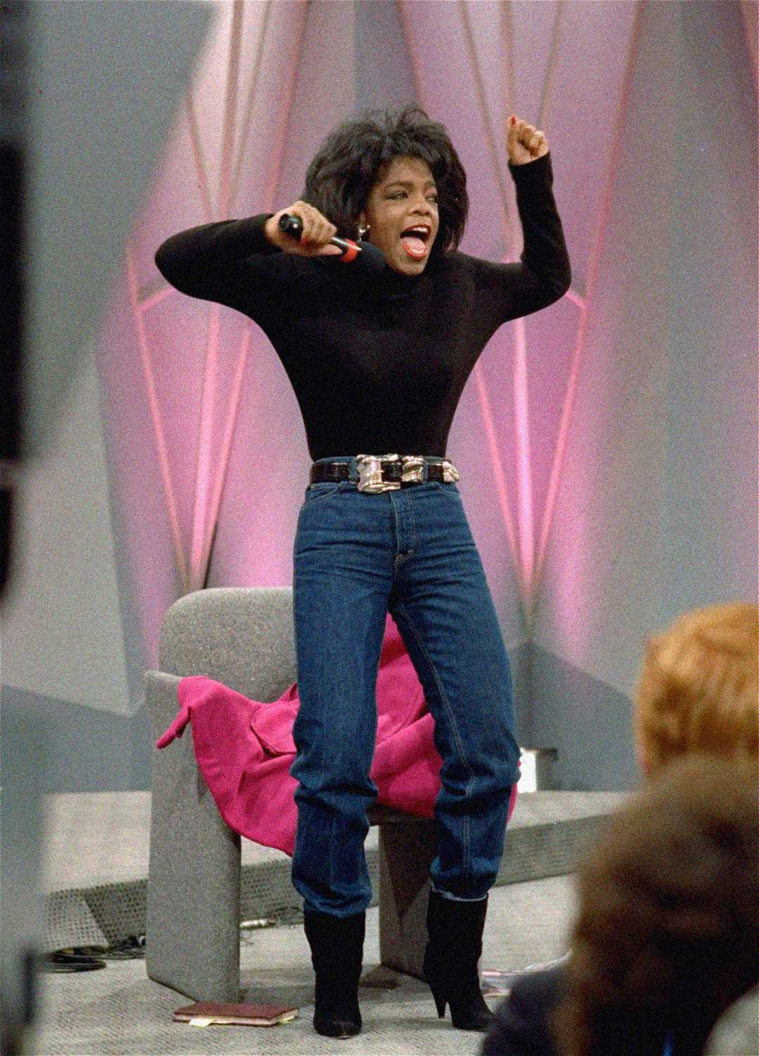 A woman in blue jeans, a black top, and black boots stands excitedly, raising her arms with a microphone in one hand. She is in front of a chair with a pink jacket draped over it, on a stage with a geometric backdrop.