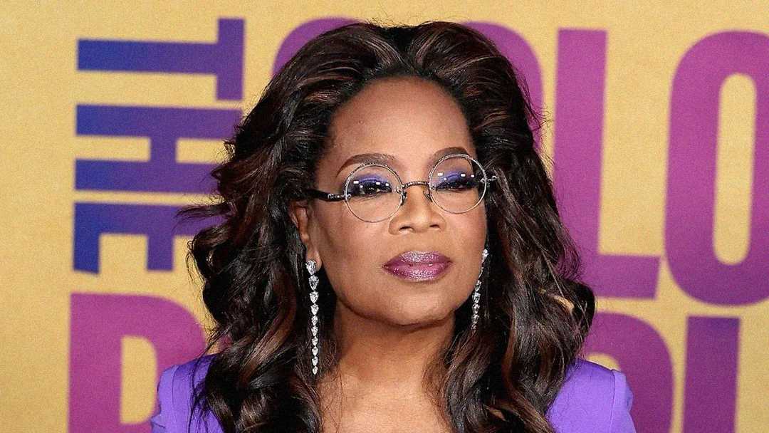 A woman with wavy brown hair and glasses, wearing long earrings and a purple outfit, stands in front of a yellow and purple background with large, partially visible text.
