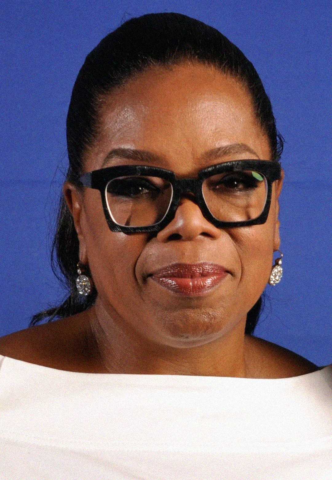 A woman with dark hair pulled back, wearing black square glasses, pearl earrings, and a white top, poses in front of a blue background.