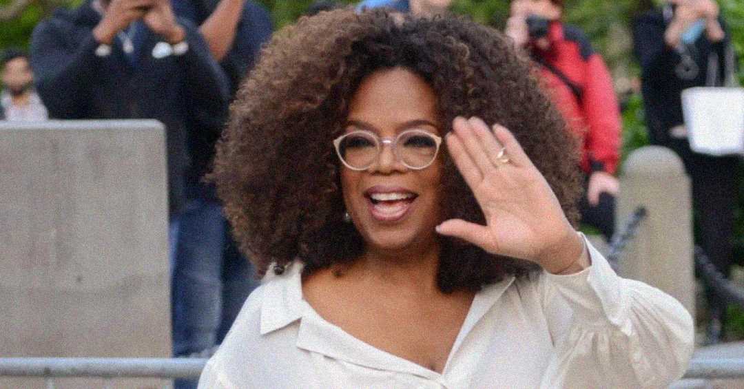 A woman with curly hair wearing glasses and a white blouse smiles and waves at the camera, with people and photographers in the background.
