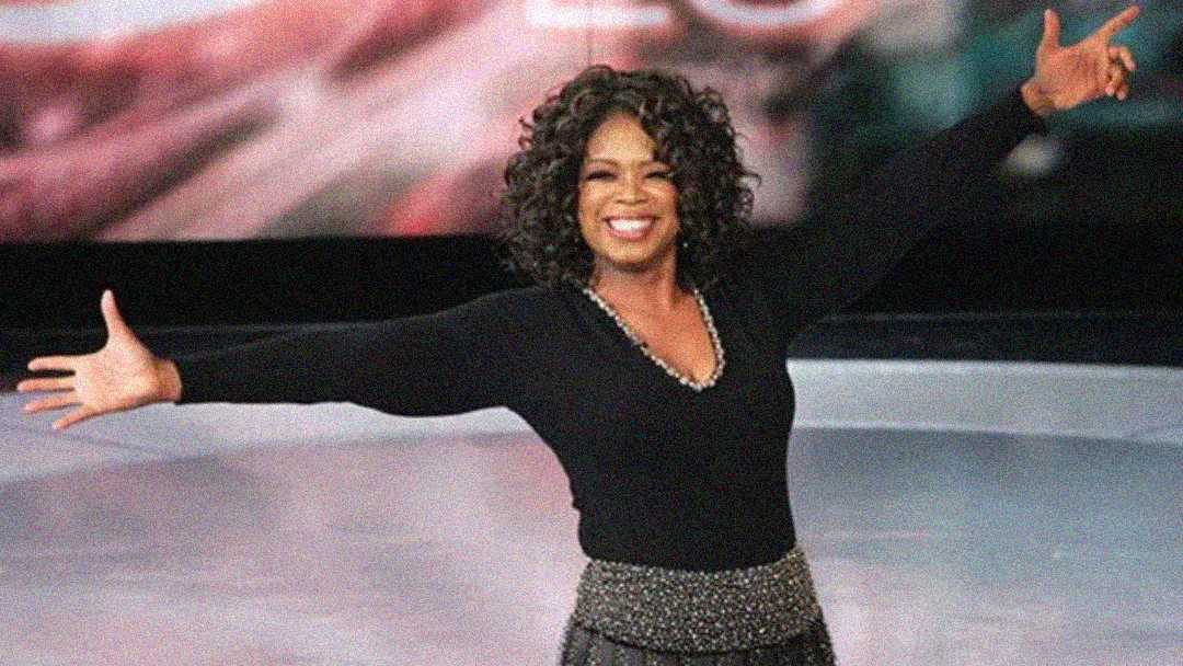 A woman with curly hair, wearing a black top and a patterned skirt, stands on a stage with her arms outstretched, smiling broadly at the audience.