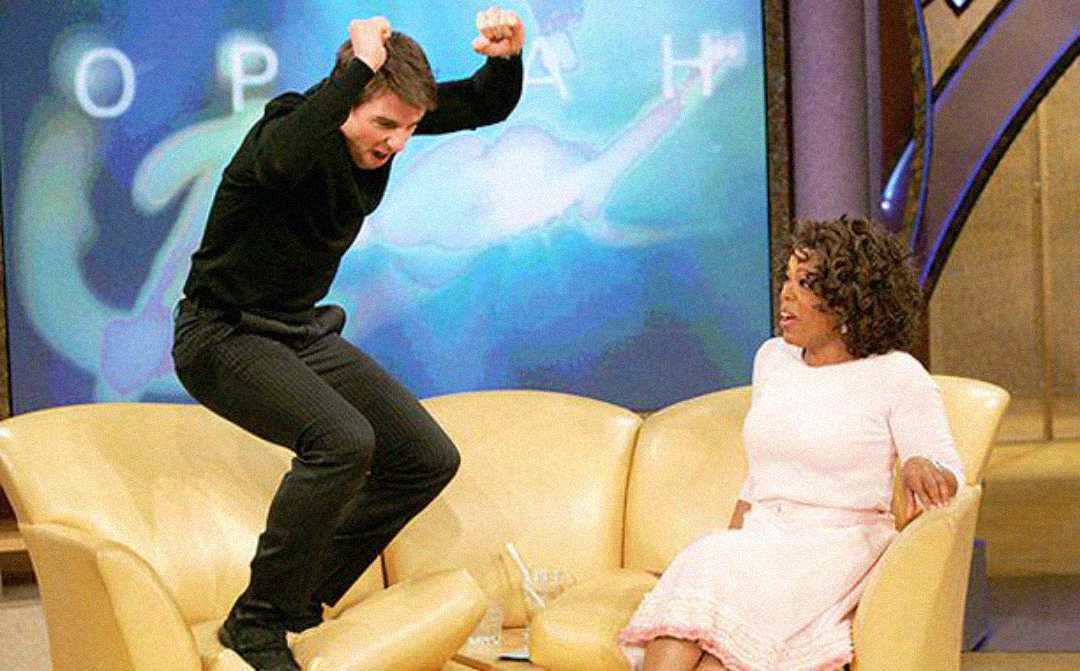 A man in black jumps excitedly on a yellow couch while a woman in a pink dress sits and looks at him surprised on a brightly lit talk show set.