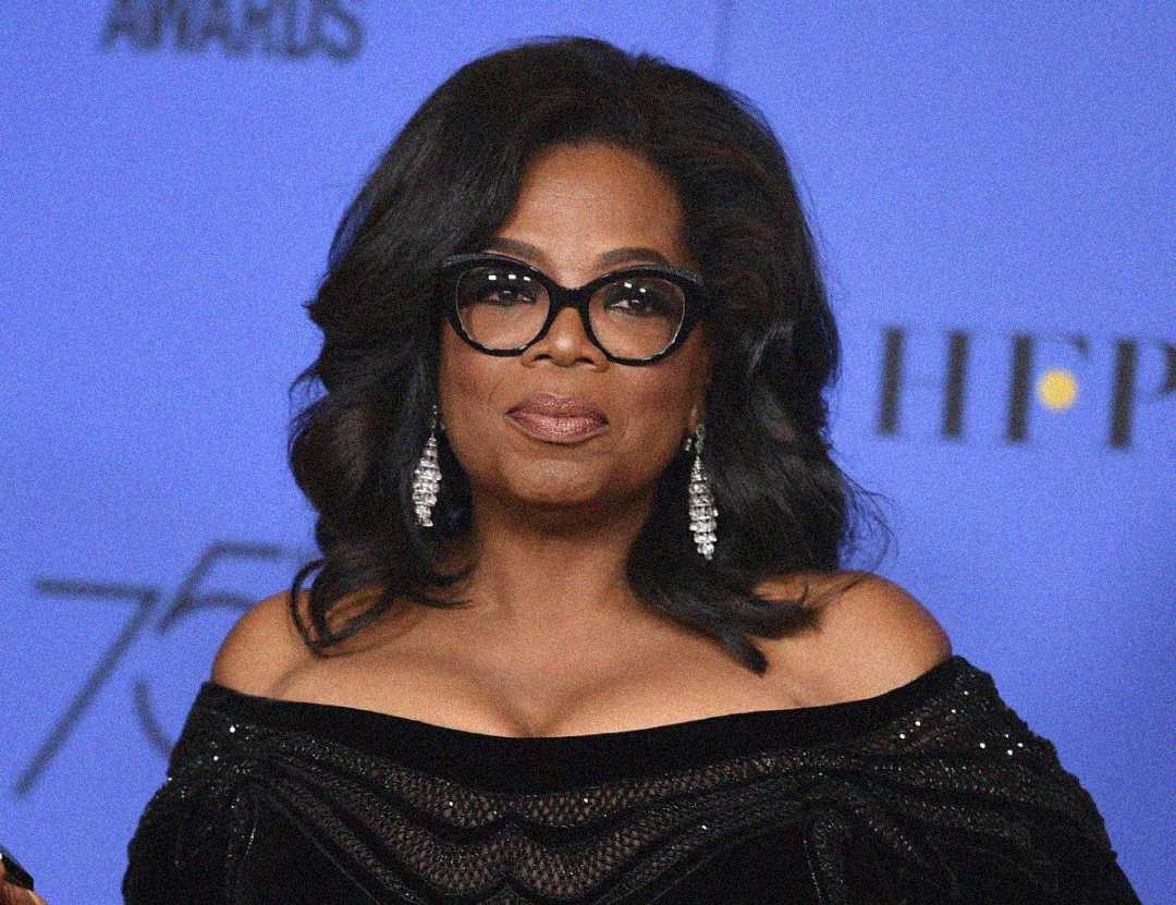 A woman with shoulder-length dark hair and glasses wears dangling earrings and a black off-the-shoulder dress, standing in front of a blue backdrop with partial text and numbers.