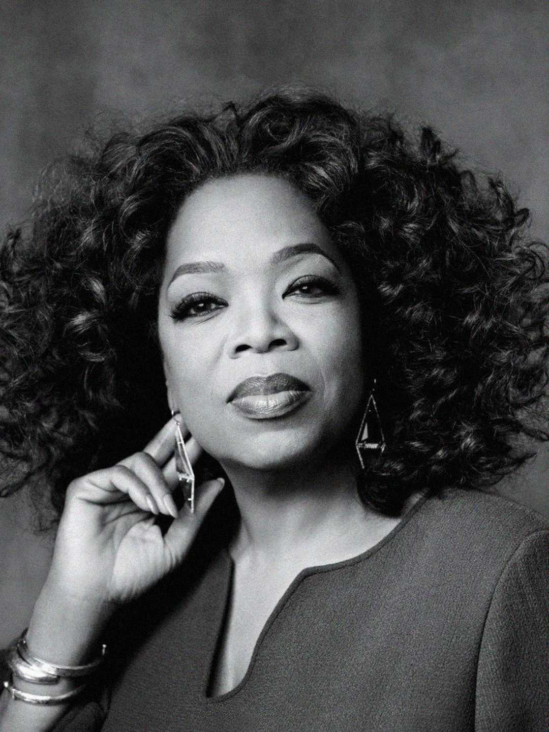 A black and white portrait of a woman with voluminous curly hair, wearing geometric earrings and a sweater. She gazes confidently at the camera with her hand touching her face.