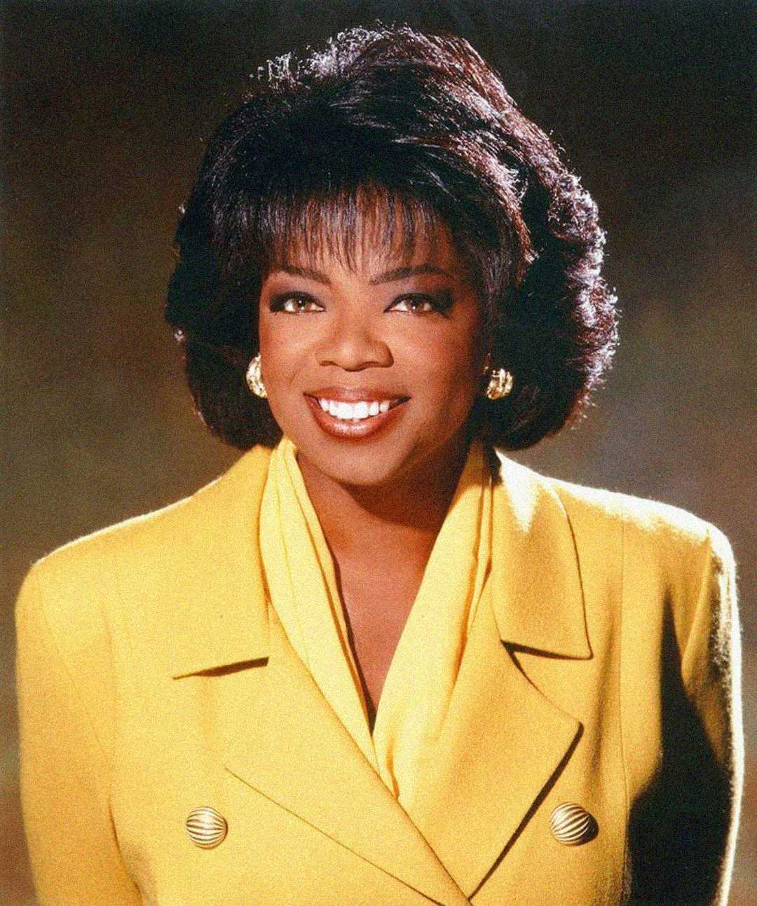 A woman with short, styled dark hair smiles warmly, wearing a bright yellow blazer with gold buttons and matching gold earrings, in front of a softly blurred brown and gold background.