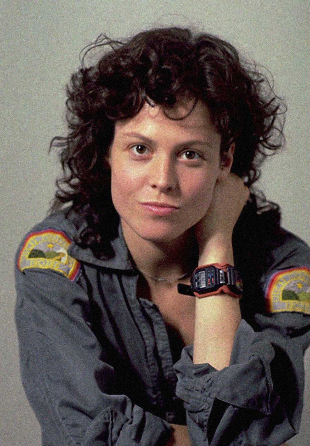 A woman with curly dark hair, wearing a gray jumpsuit with colorful patches on the shoulders and a digital wristwatch, sits with her head resting on her hand, looking confidently at the camera.