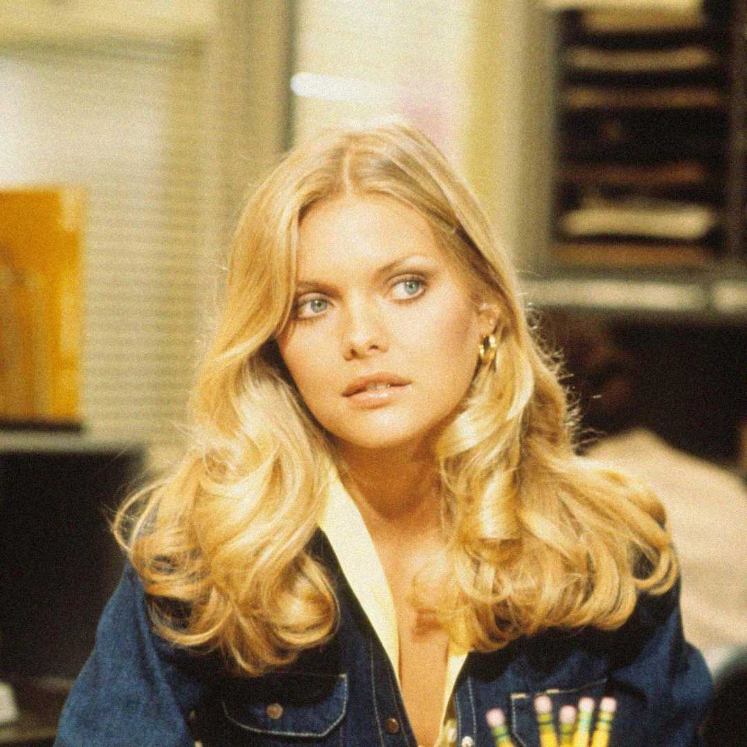 A woman with long, wavy blonde hair and light eyes sits indoors, wearing a dark jacket over a light blouse. She looks slightly to the side with a neutral expression. Office supplies and shelves are visible in the background.