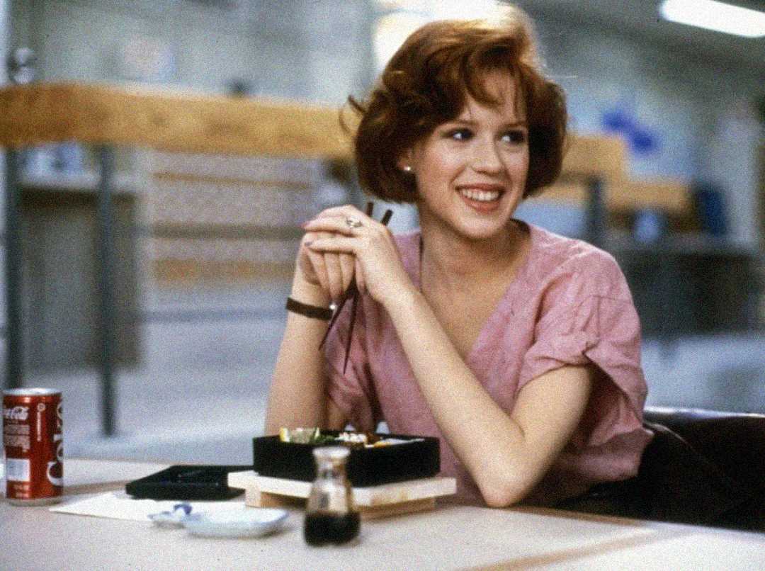 A young woman with short red hair wearing a pink blouse sits at a table, smiling. In front of her are a food container, a soda can, chopsticks, and a small bottle of soy sauce. The background is a blurred indoor space.