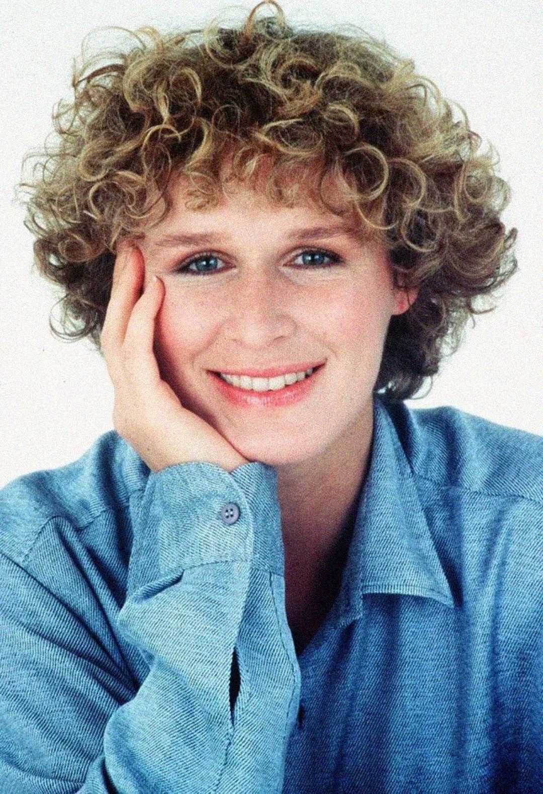 A person with short, curly blonde hair and blue eyes smiles at the camera, resting their face on their hand. They are wearing a light blue collared shirt against a plain white background.