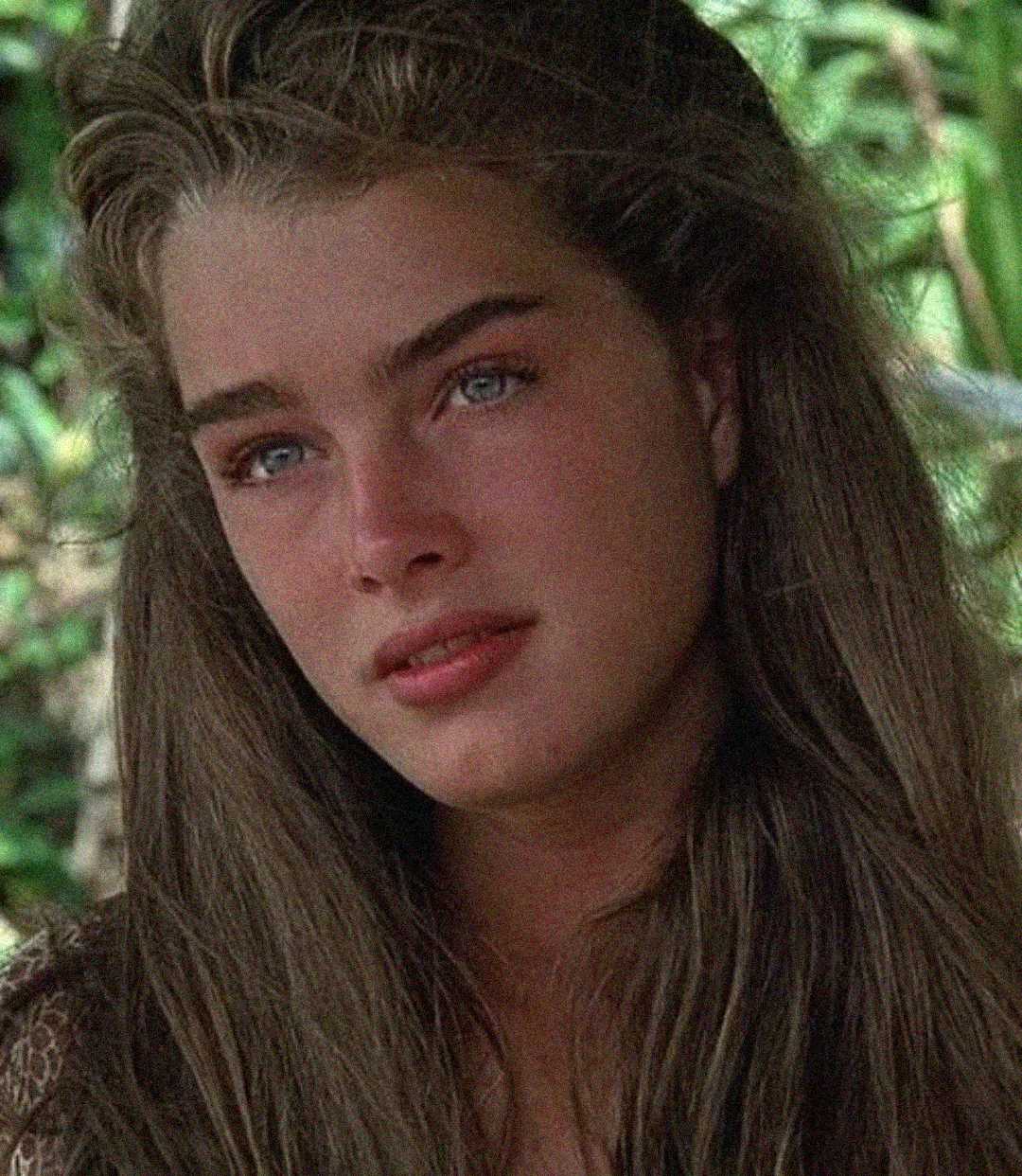 A young woman with long brown hair and blue eyes gazes slightly to the side, surrounded by greenery. She has natural makeup and a calm, thoughtful expression.