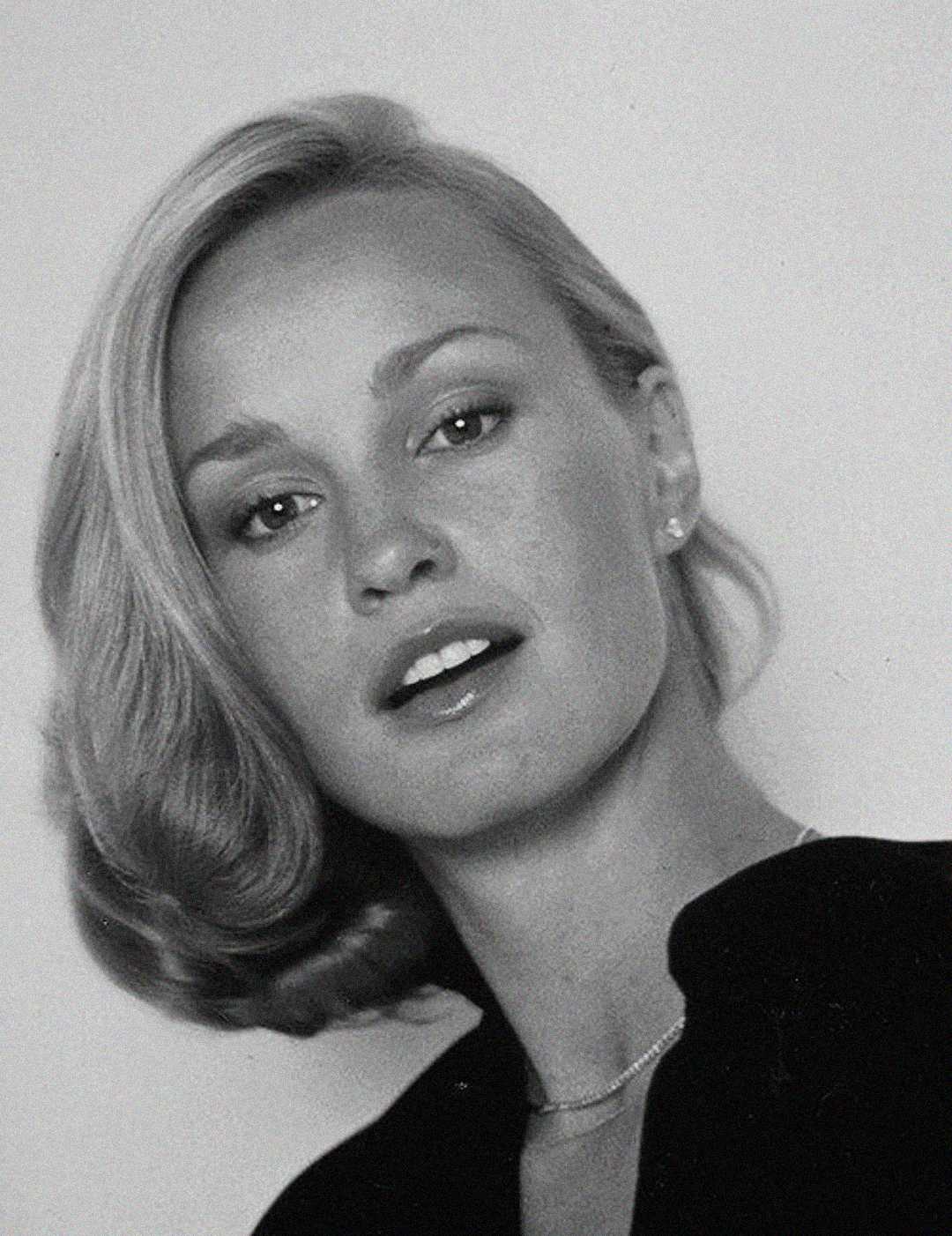 Black and white portrait of a woman with light hair styled in waves, wearing a dark top and a necklace, looking slightly up and away from the camera with a neutral expression.