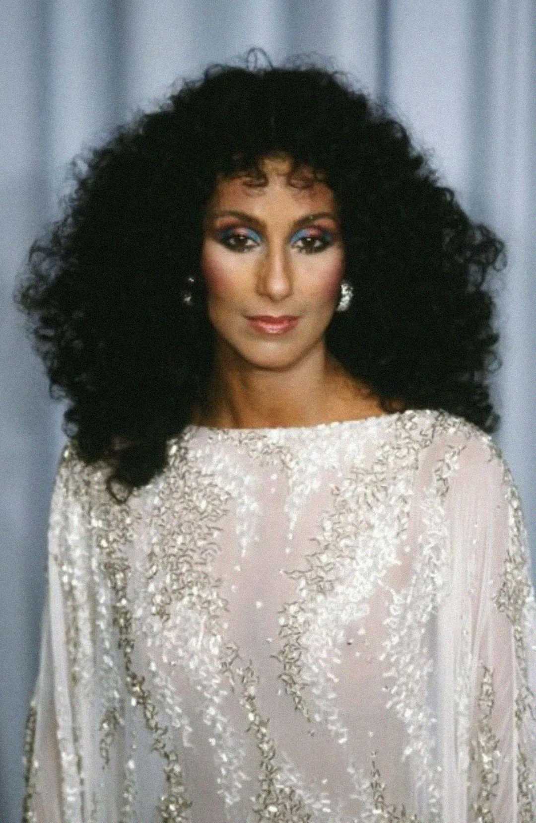 A woman with voluminous curly dark hair wears a sparkling, intricately beaded white gown and large earrings, standing in front of a light gray background. She has dramatic makeup and a composed expression.