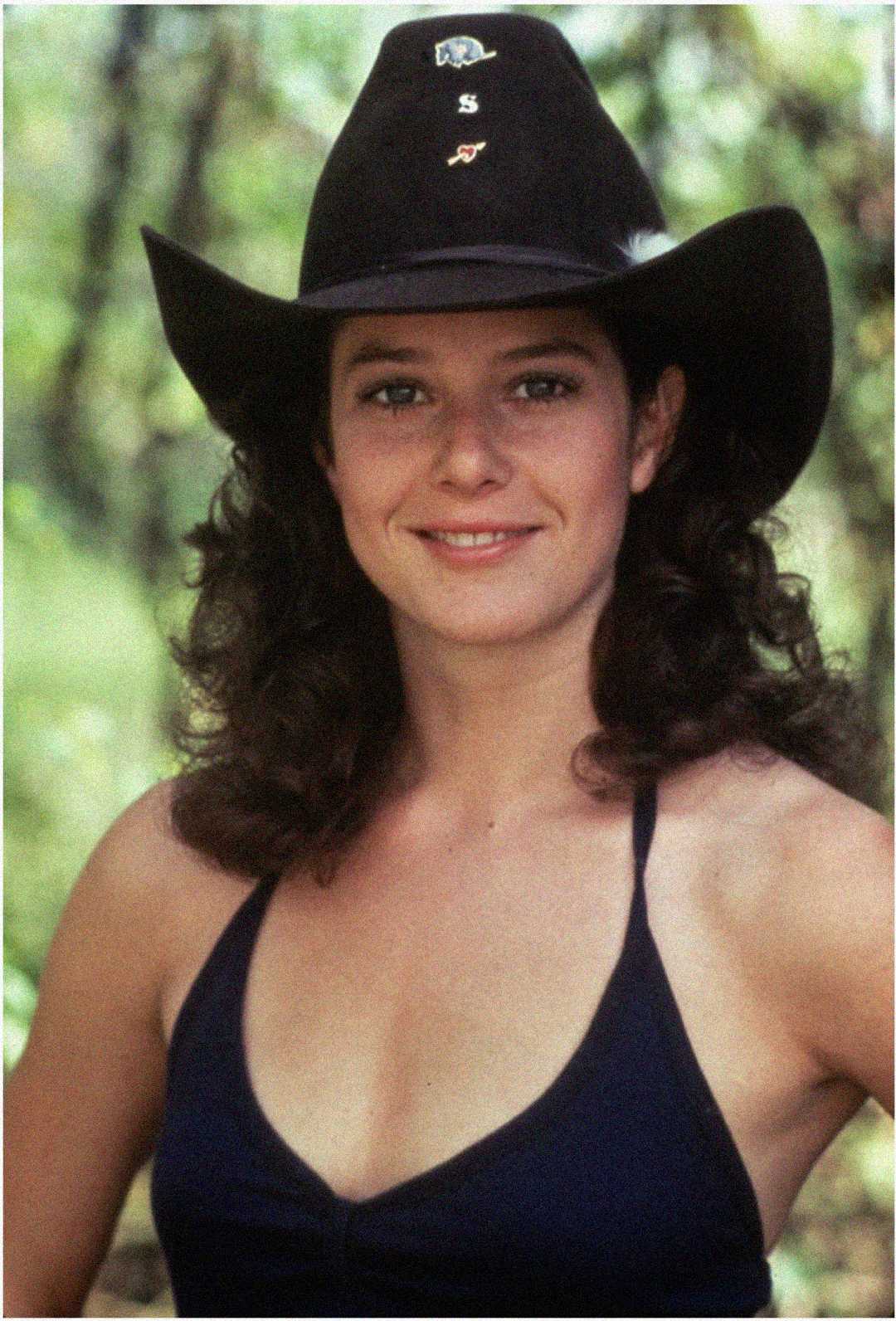 A woman with dark curly hair wearing a black cowboy hat and a dark halter top smiles outdoors, with greenery blurred in the background.