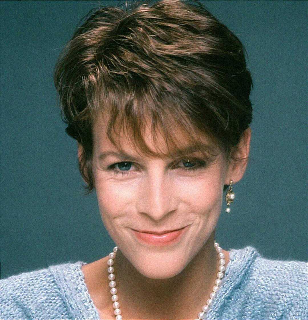 A person with short brown hair, wearing a light blue sweater, a pearl necklace, and a dangling earring, smiles softly against a plain grey background.