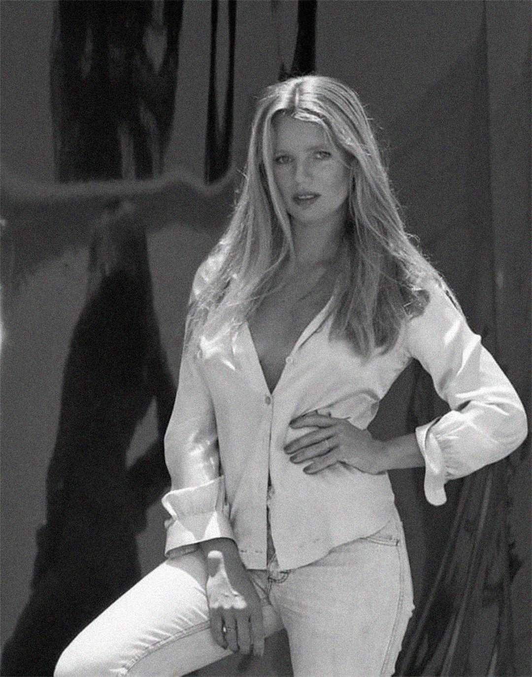 A woman with long, light hair poses confidently in a partially unbuttoned shirt and light jeans. She stands with one hand on her hip against a dark, reflective background. The image is black and white.
