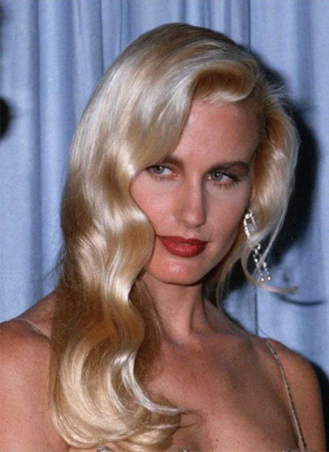 A woman with long, wavy platinum blonde hair, red lipstick, and blue eyes poses in front of a light blue curtain. She is wearing a spaghetti strap top and dangling earrings.