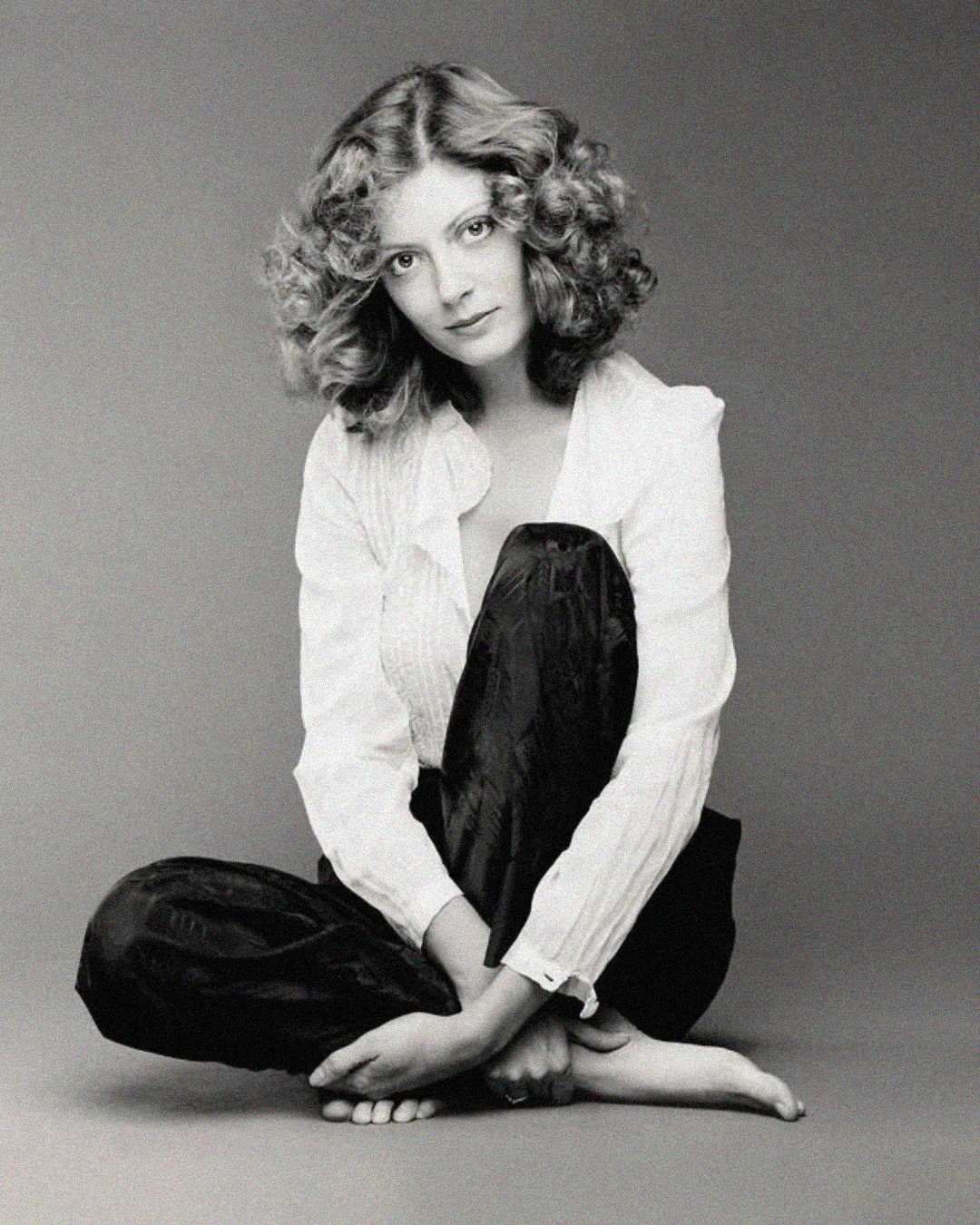 A woman with curly hair sits barefoot on the floor, knees up and arms wrapped around her legs. She wears a white shirt and dark pants, looking softly at the camera against a plain background. The image is in black and white.