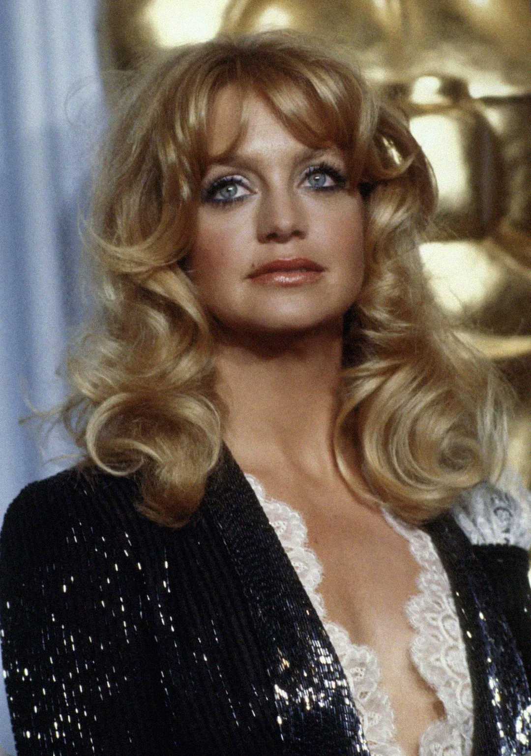 A woman with long, wavy blonde hair and blue eyes wears a black sequined jacket over a white lace top, standing in front of a gold background.