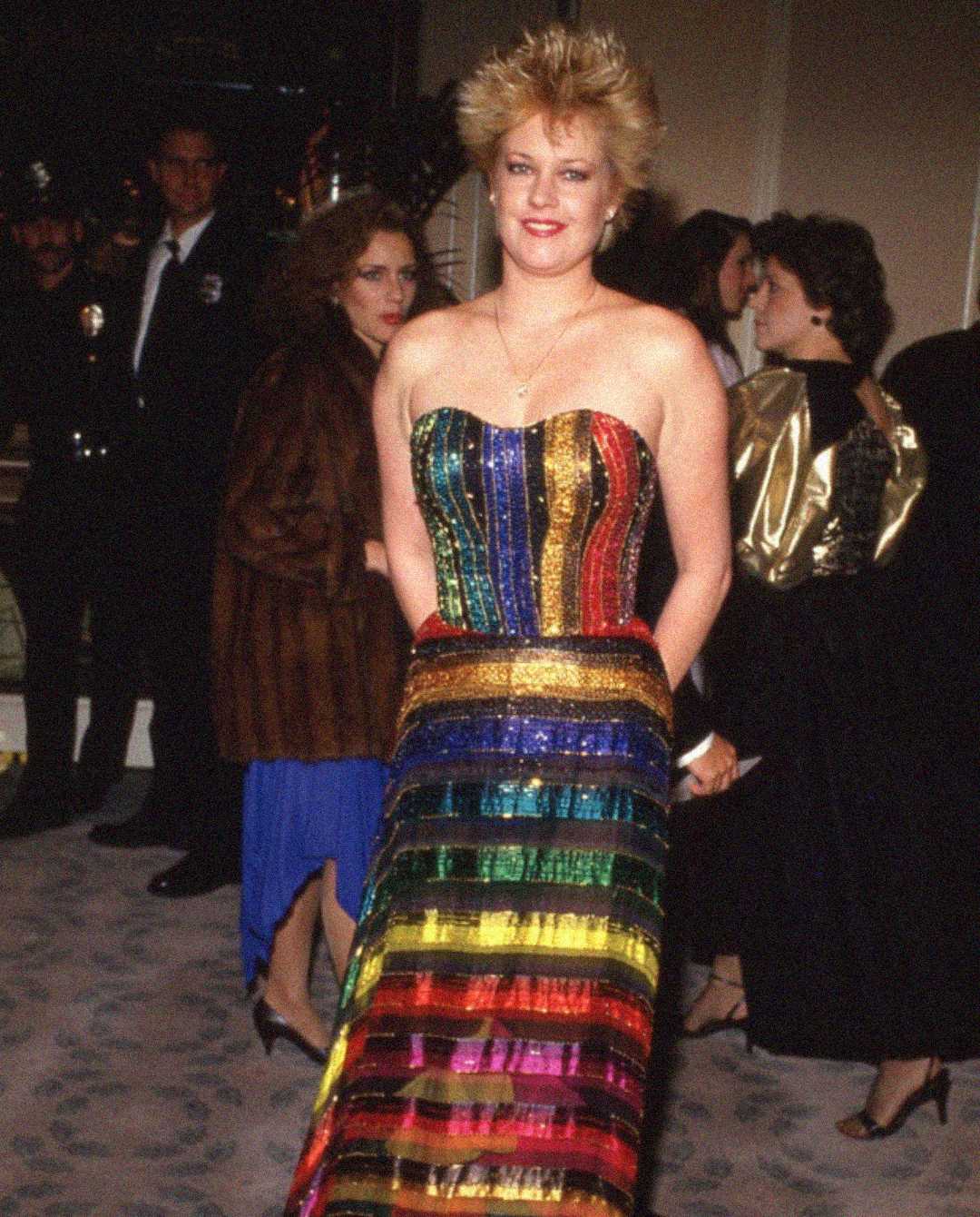 A woman in a strapless, rainbow-striped, sequined dress smiles at an indoor event. Other guests in formal attire and a police officer are visible in the background.