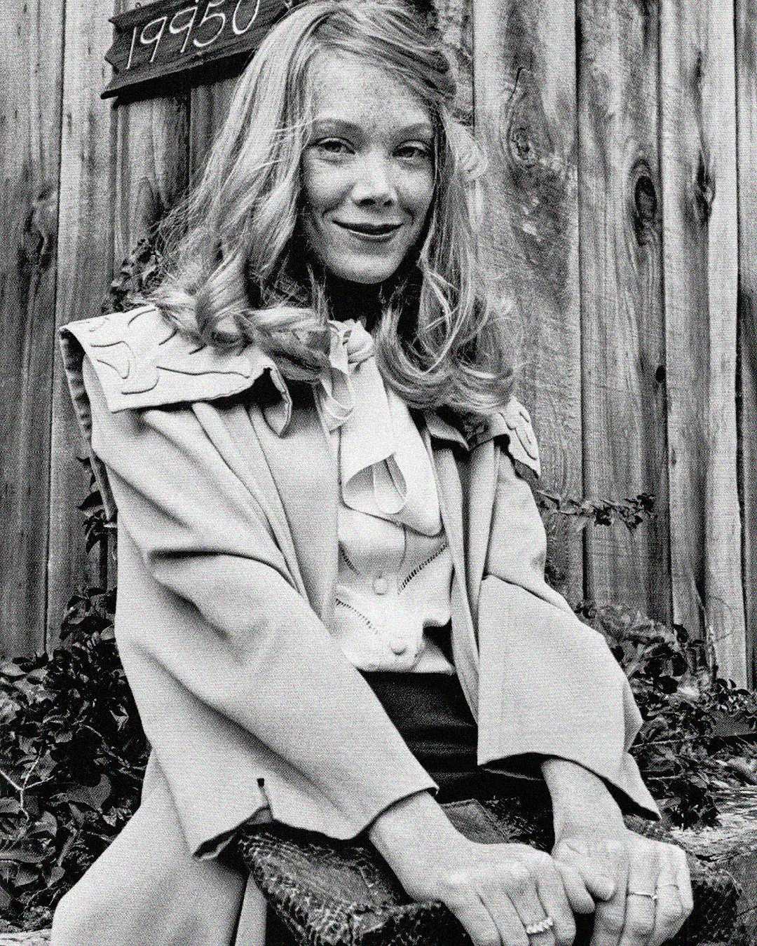 A young woman with long, wavy hair and freckles smiles while sitting in front of a wooden fence. She wears a light-colored jacket over a blouse with a bow and has her hands resting on a textured object.