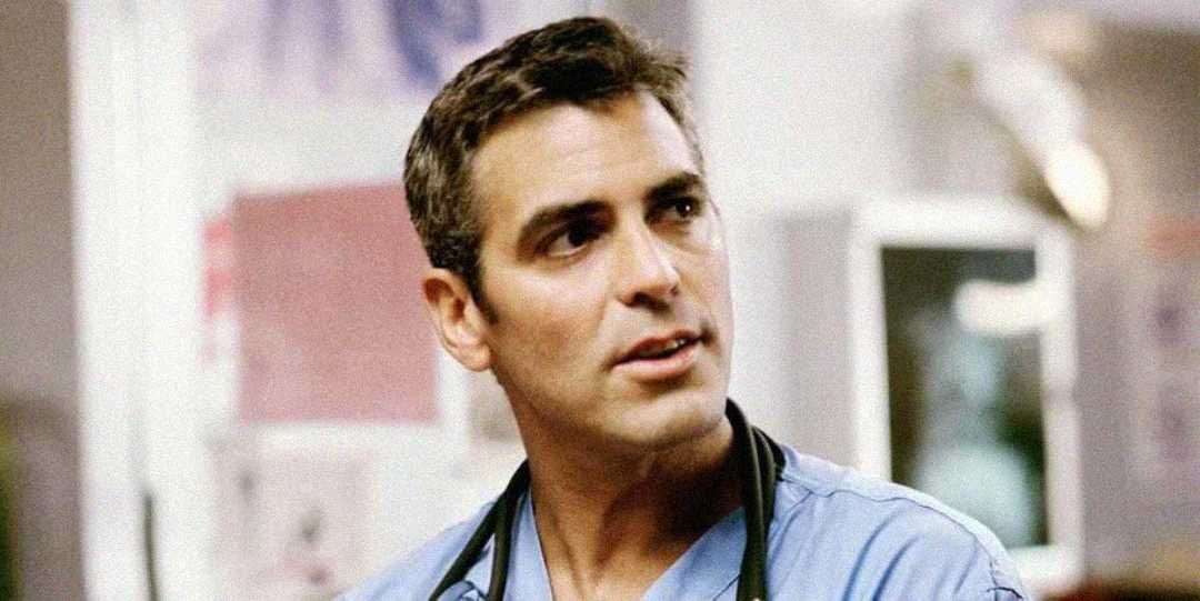 A man wearing blue medical scrubs and a stethoscope around his neck stands indoors, looking slightly to the side. The background is blurred, suggesting a hospital or clinical setting.