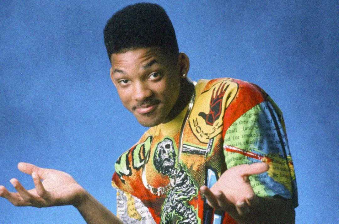 A young man with a flat-top haircut shrugs with both palms up, wearing a colorful, patterned shirt against a blue background. He is smiling slightly and tilting his head.