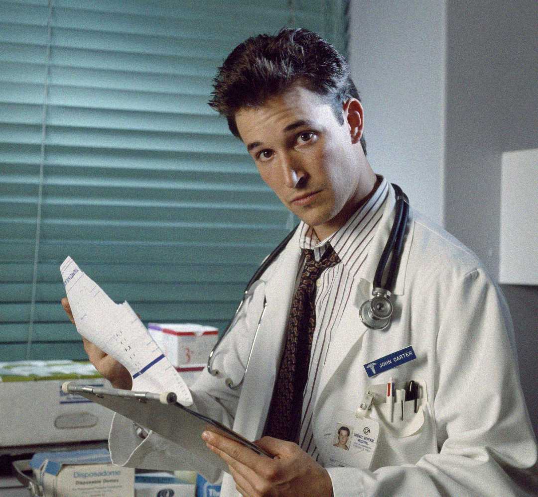 A young male doctor with a stethoscope around his neck holds a clipboard and a torn sheet of paper, standing in a medical office with blinds and supplies in the background.