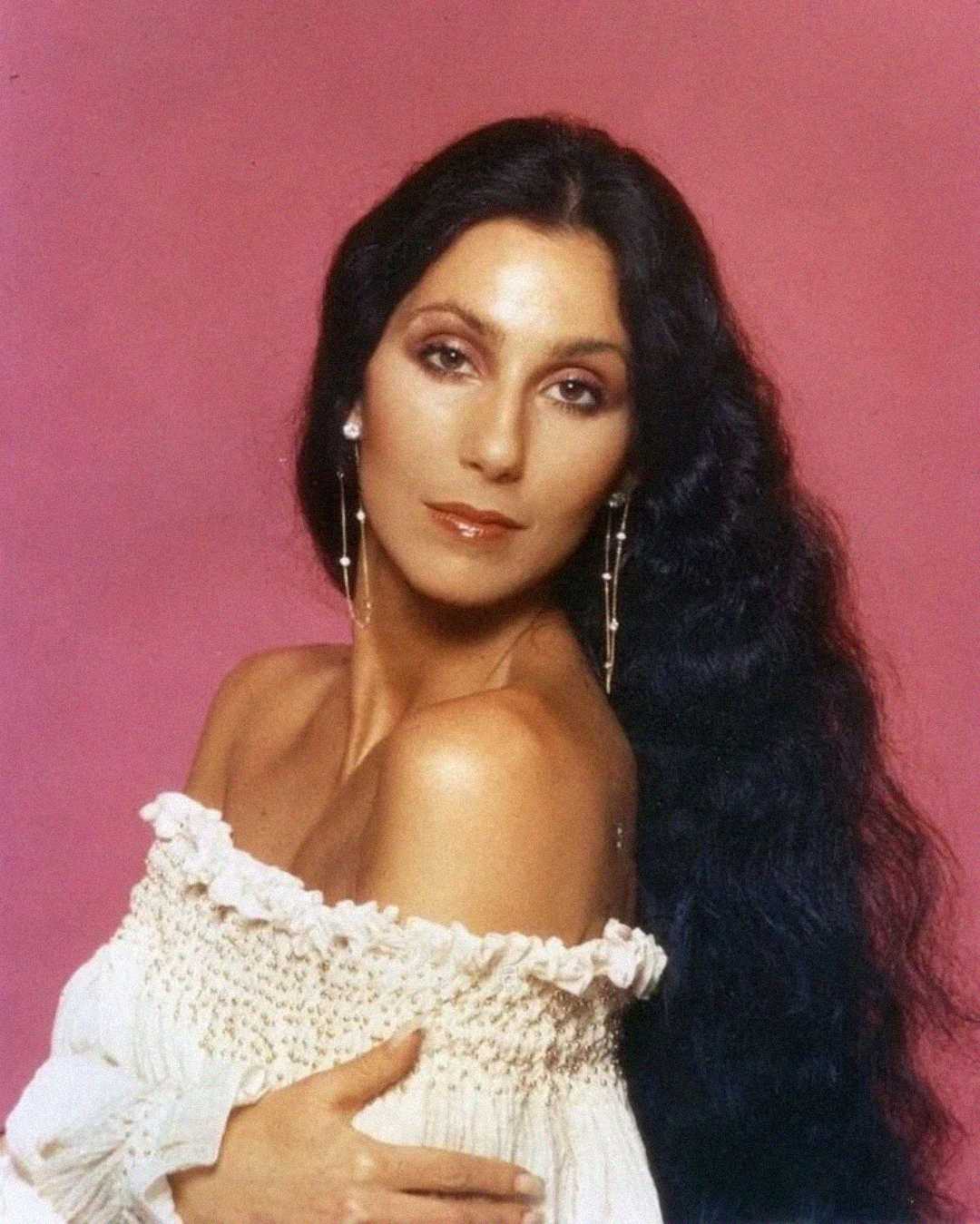 A woman with long black hair and dangling earrings poses against a pink background, wearing an off-the-shoulder white top and gazing softly at the camera.