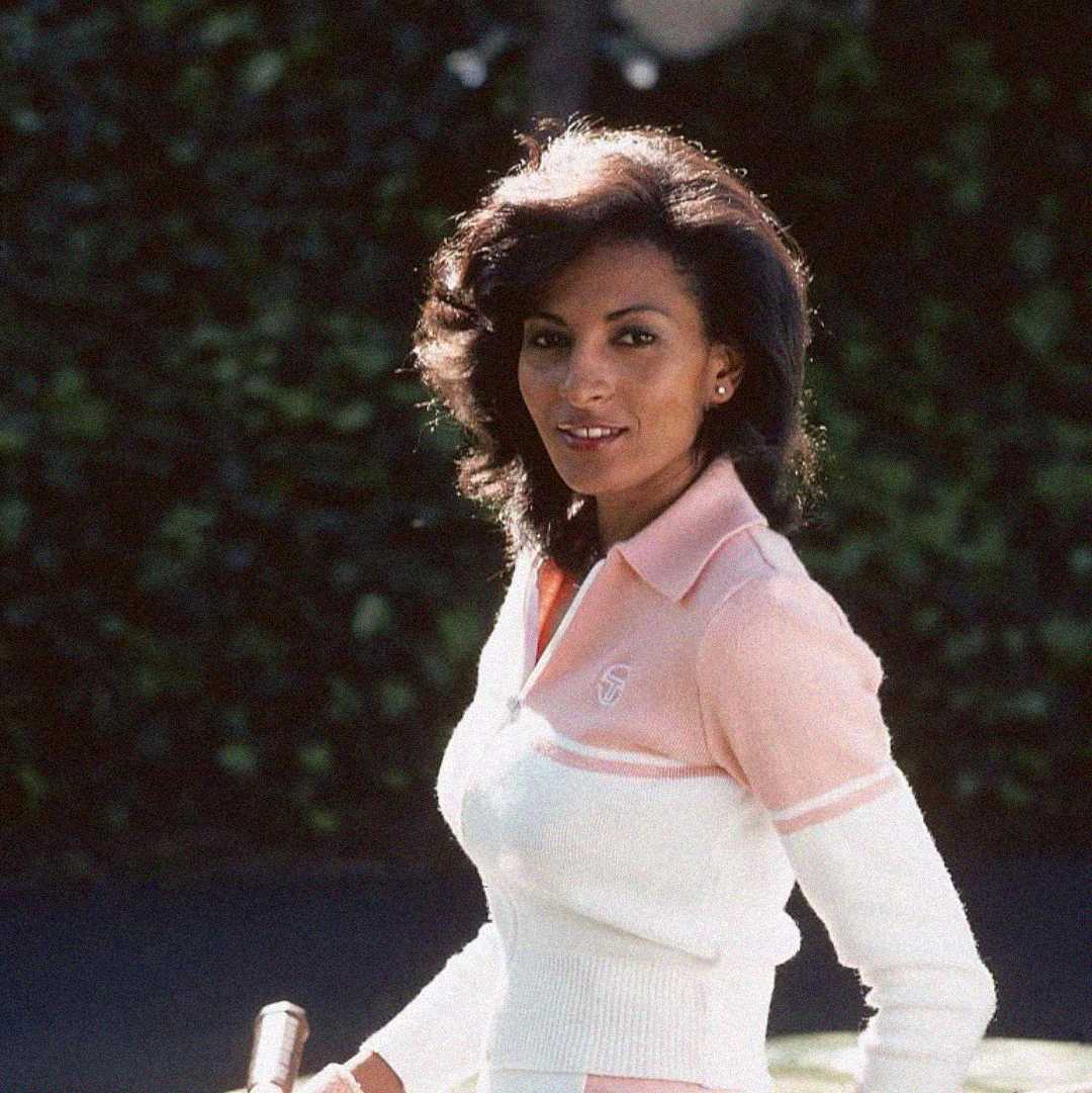 A woman with shoulder-length dark hair wearing a white and pink collared sweater stands outdoors, holding a tennis racket, with green foliage in the background.