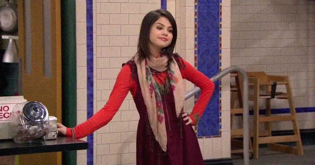 A young woman with shoulder-length dark hair stands confidently indoors, wearing a red long-sleeve top, a maroon dress, and a beige patterned scarf. She has one hand on her hip and is leaning on a counter.