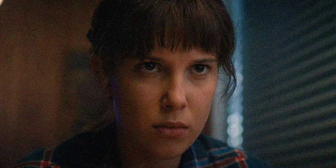 A young person with straight brown hair and bangs stares intently forward with a serious expression, wearing a plaid shirt. The background is dimly lit with horizontal blinds visible on the right side.