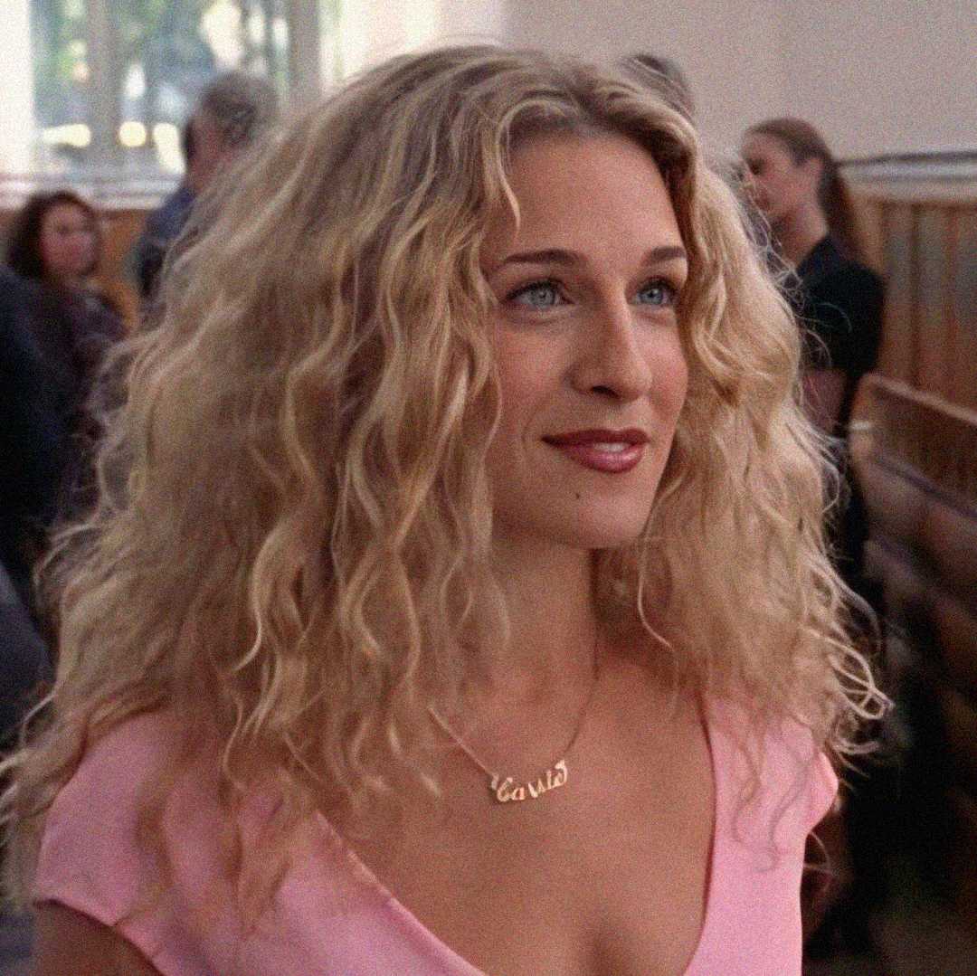 A woman with long, curly blonde hair and blue eyes is wearing a pink top and a gold necklace that says "Carrie." She is smiling slightly and standing indoors, with people blurred in the background.