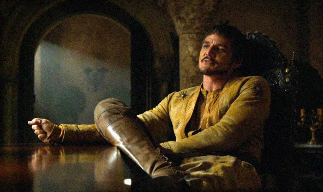A man in a yellow tunic sits relaxed in a dimly lit room, leaning back in a chair with one arm resting on a table and his leg propped up. Sunlight streams through a window behind him, creating a dramatic atmosphere.