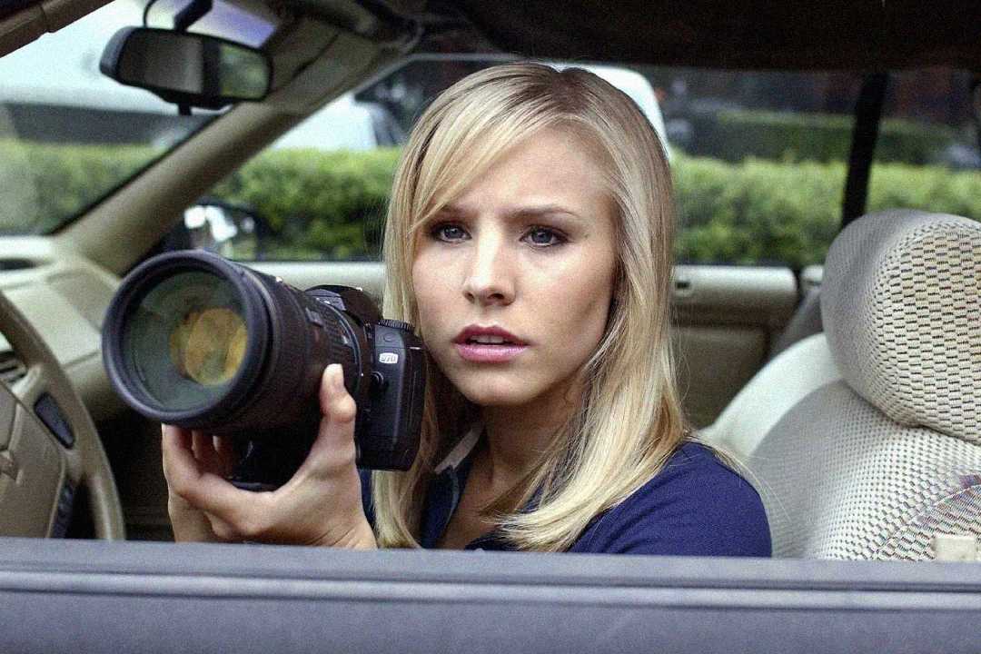 A woman with blonde hair is sitting in a car, holding a camera with a large lens, looking intently out the window as if observing or photographing something.