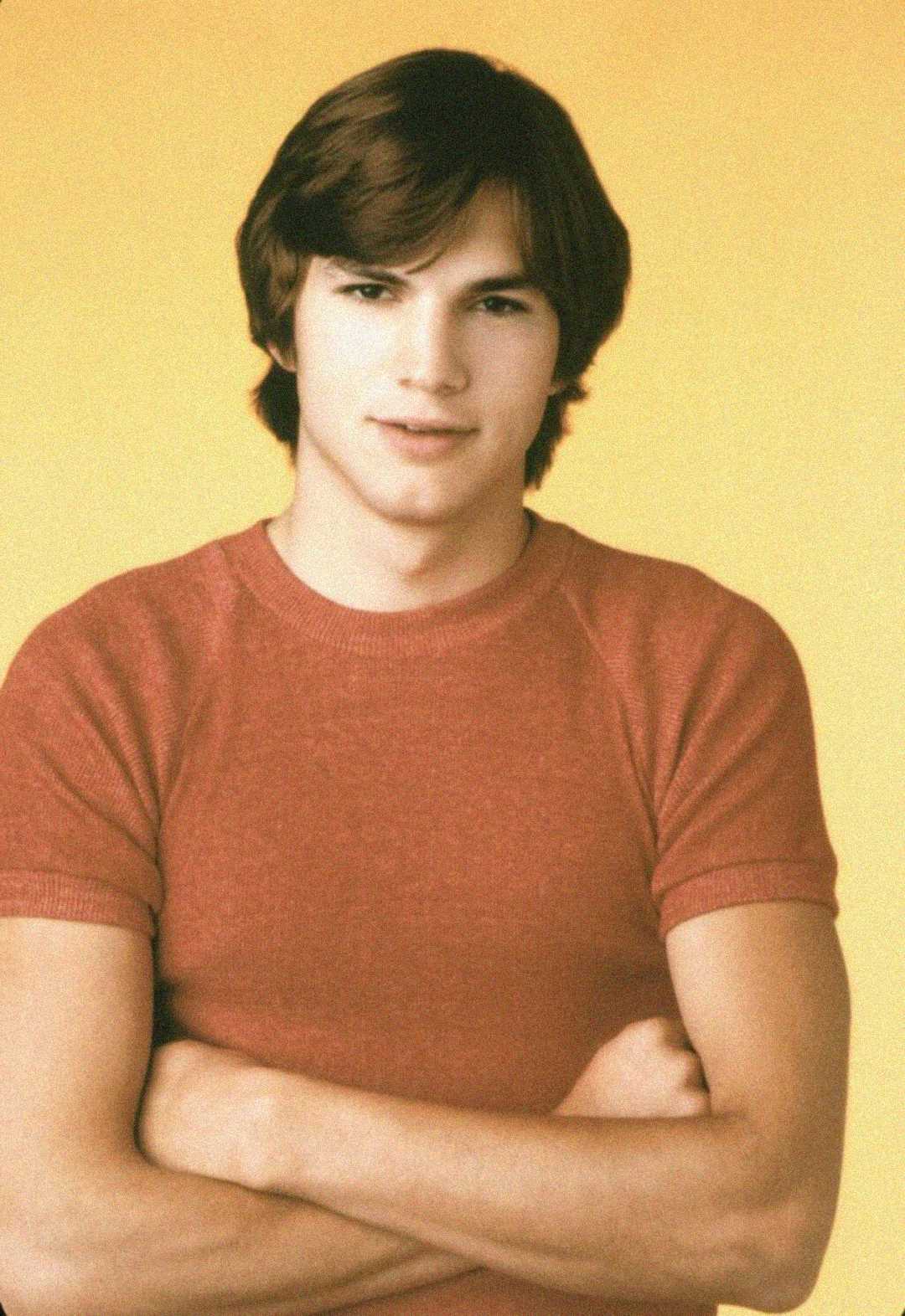 A young man with medium-length brown hair stands against a yellow background, wearing a short-sleeved red shirt and crossing his arms with a neutral expression.