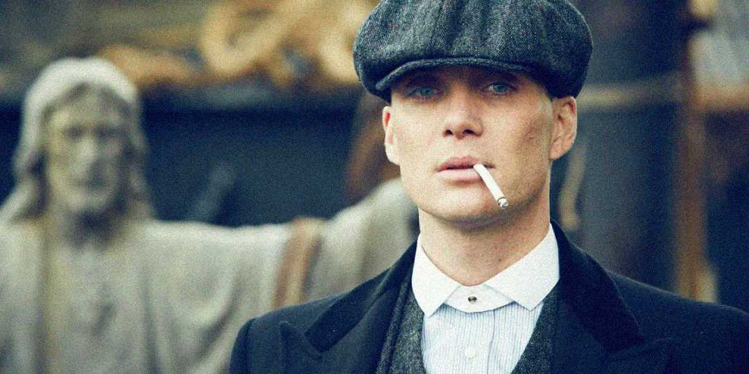 A man wearing a flat cap and dark coat looks ahead with a serious expression, a cigarette hanging from his lips. A blurred statue with outstretched arms appears in the background.