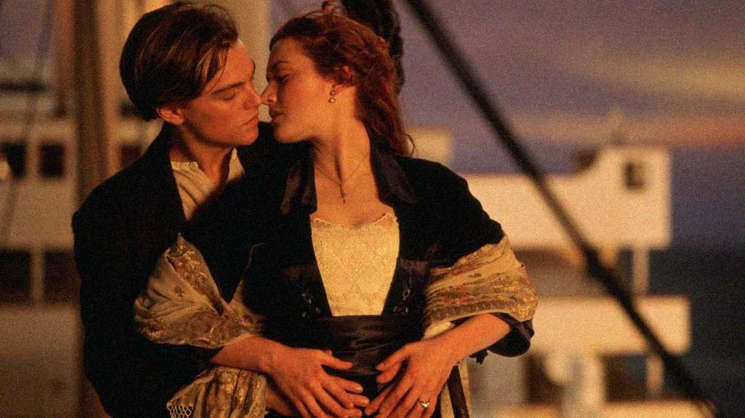 A young couple stands close together on a ship's deck at sunset, the man holding the woman around her waist as they gaze lovingly into each other's eyes.